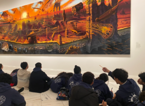 Ms. Fisher's 8th grade students sit on the gallery floor, looking up and gesturing towards the large painting on the wall (Manifest Destiny).